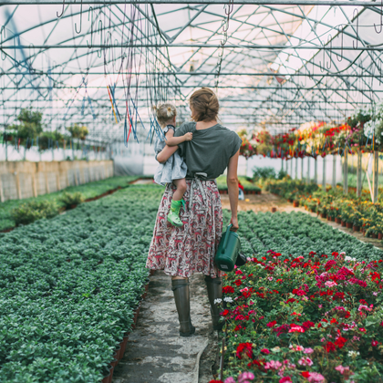 woman in skirt holding child on her left hip, standing in large community garden holding a watering can