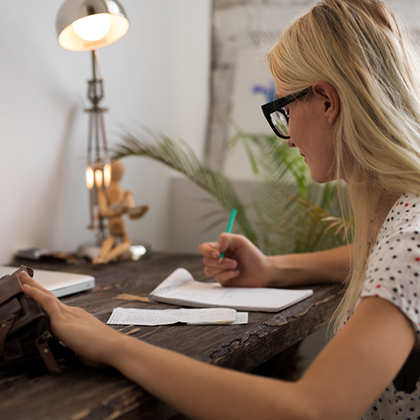 woman with long blond hair wearing glasses and a white polka dotted short-sleeved top, sitting at a desk writing on a notepad