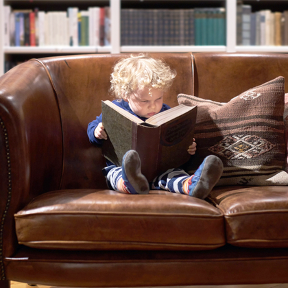 toddler reading large book on brown leather couch