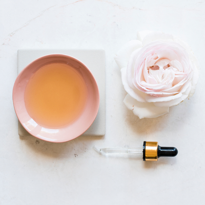 rose water in a pink cup, a light pink rose and a Pasteur pipette laid out on white surface