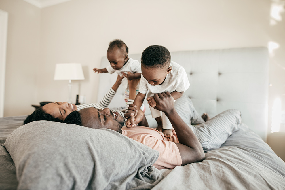 A young family lays together in bed on a sunny morning; the parents are lifting up their children and playing with them.