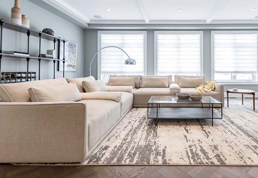 A big white couch in front of a white marble fire place in a room with big windows and a silver lamp 