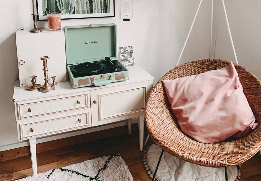 A basket chair with a pink pillow next to a white cabin and a record player