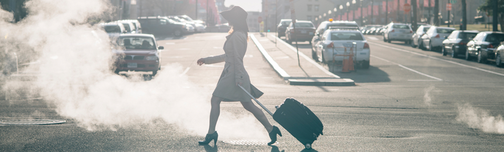 girl in heels, dress and hat crossing street pulling suitcase