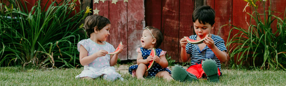 three toddlers sitting outside in the grass eating watermelon